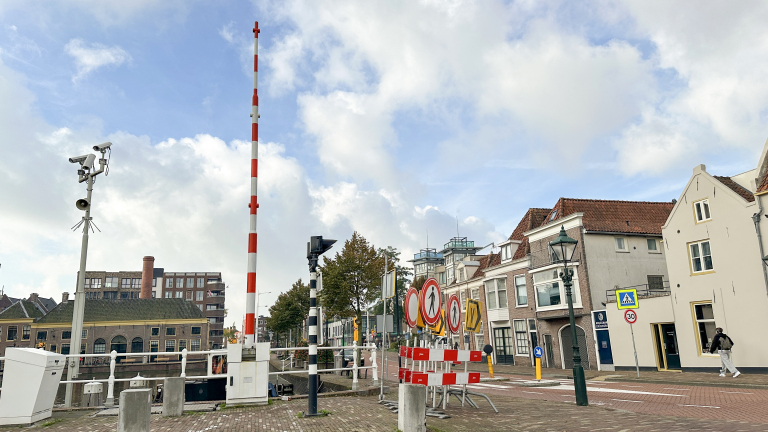 Rood-wit slagboom bij een brug met verkeersborden en oude huizen langs een kanaal onder een bewolkte lucht