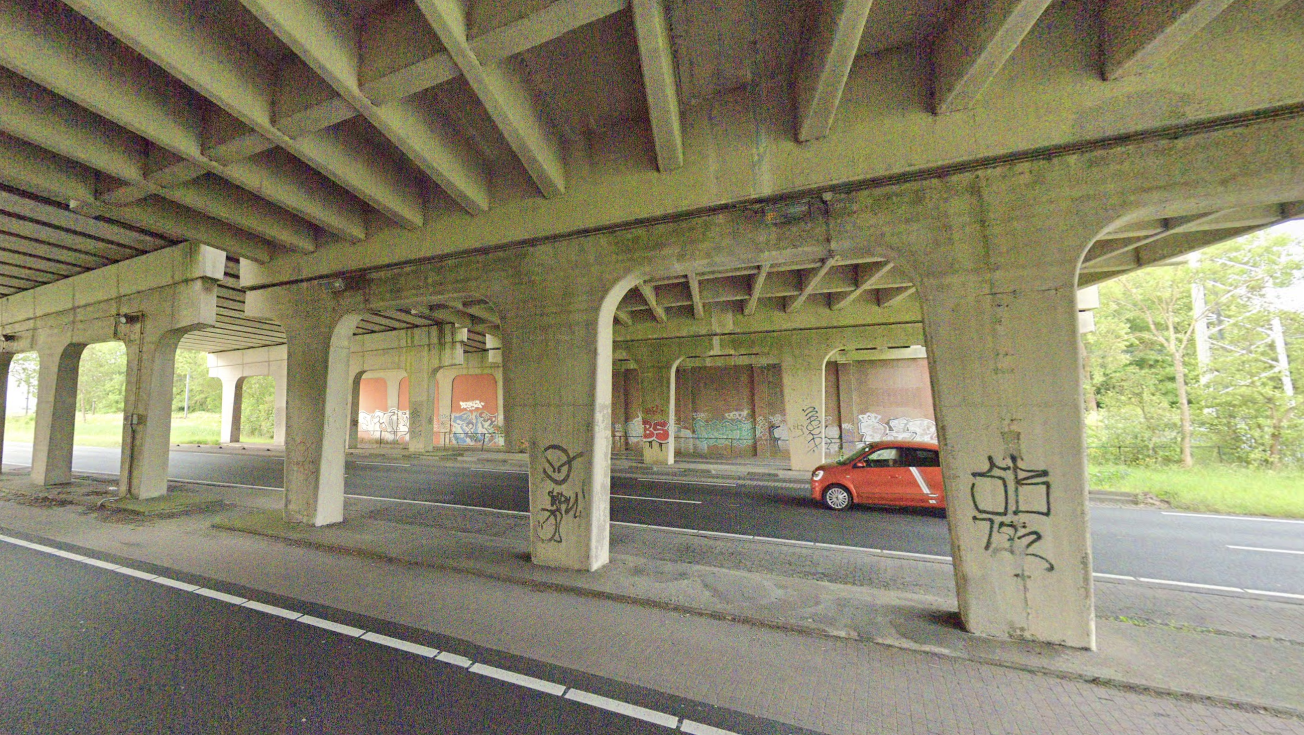 Onder een betonnen viaduct met graffiti op pijlers, met een rood autootje op de weg en bomen op de achtergrond
