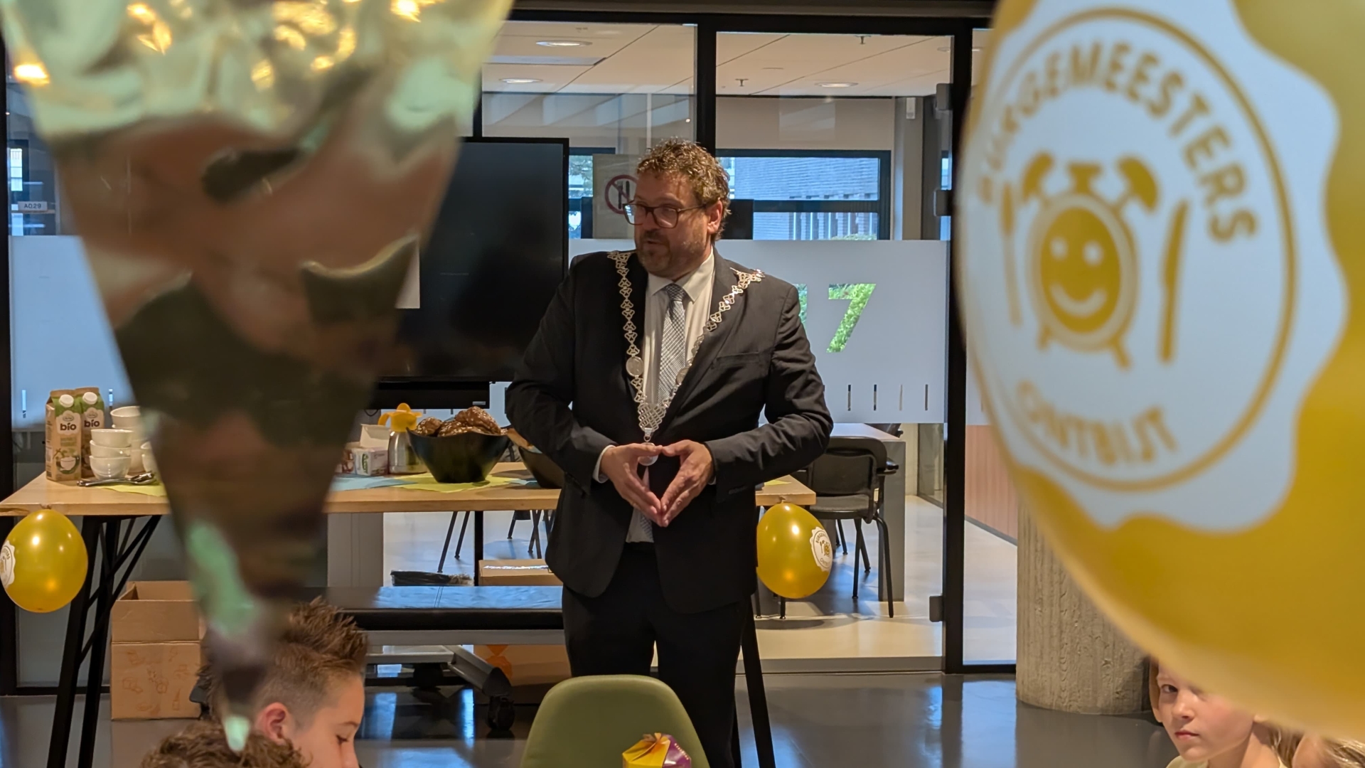 Man in pak met ambtsketen spreekt in een ruimte met tafels en gele ballonnen met logo en tekst “meester”