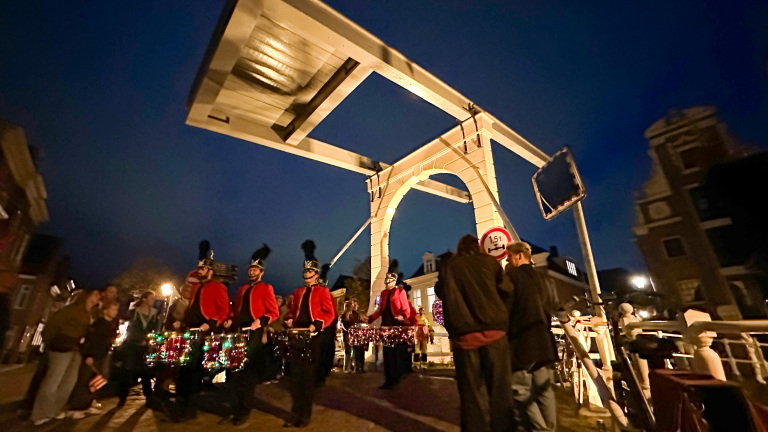 Drumband in rode uniformen marcheert ’s avonds onder een openstaande ophaalbrug, met publiek eromheen