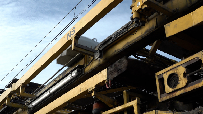 Gele NS-railwerkmachine met bewegende onderdelen onder bovenleidingsdraden tegen een lichte lucht