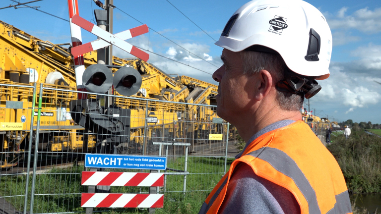 Spoorwerker met witte helm en oranje veiligheidshesje bij spoorwegovergang met bord WACHT en gele werkmachines