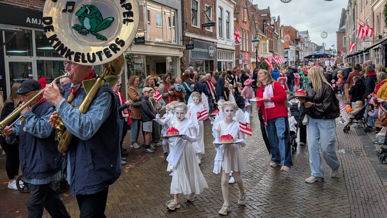 Optocht in winkelstraat met fanfare en kinderen in witte outfits die taarten dragen en rood-witte vlaggetjes zwaaien
