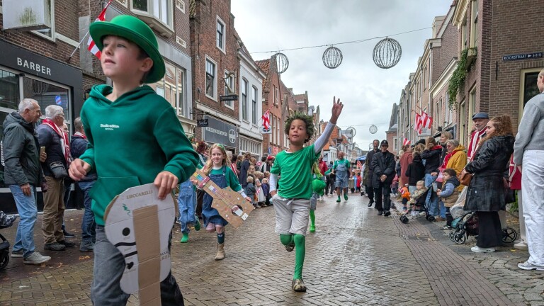 Kinderen in groen lopen in een optocht door een straat, met toeschouwers langs de kant en rood-witte vlaggen