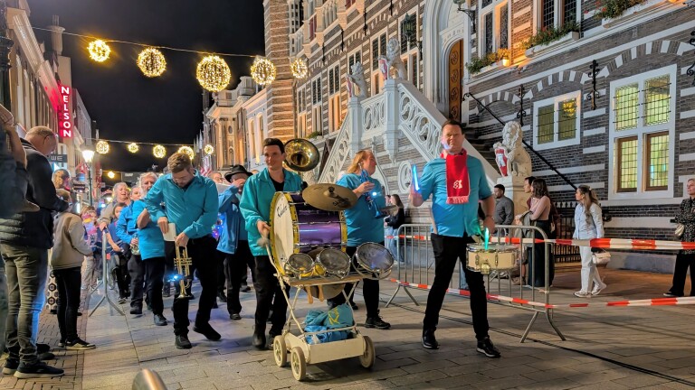 Fanfare loopt door verlichte straat bij nacht langs publiek, met trommels op karretje voor historisch gebouw