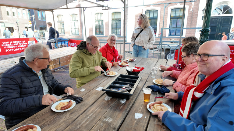 Groep mensen eet aan lange picknicktafel onder een tent, met borden rookworst en stamppot op een plein