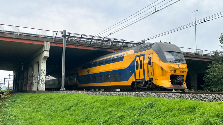 Gele en blauwe dubbeldekker NS-trein rijdt onder een viaduct langs groen gras en bovenleidingen