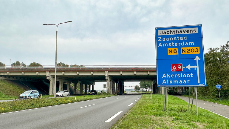 Blauw verkeersbord bij viaduct met verwijzingen naar Zaandstad, Amsterdam (N8/N203) en A9 naar Akersloot en Alkmaar