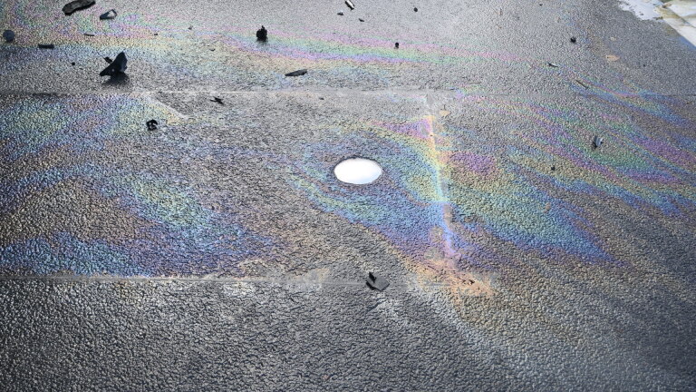 Olievlek met regenboogkleurige glans op nat asfalt, met verspreide brokstukken en een ronde putdeksel
