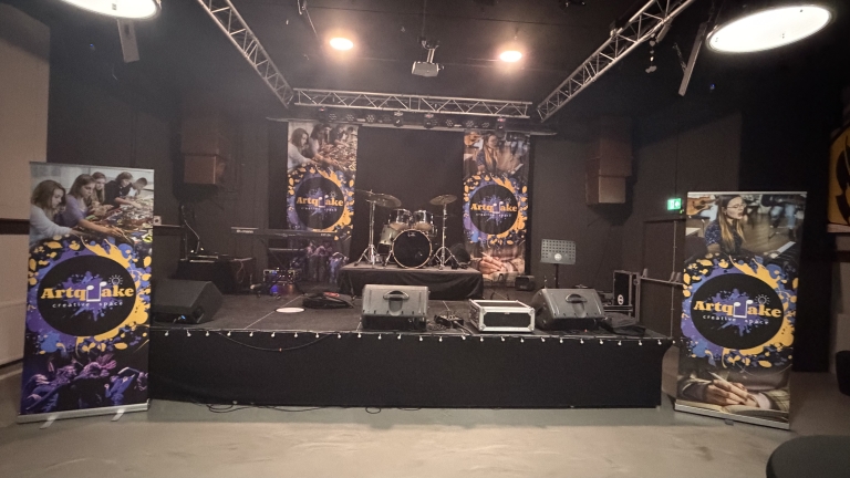 Podium met drumstel, speakers en Artquake creative space-banners in een donkere zaal met podiumverlichting