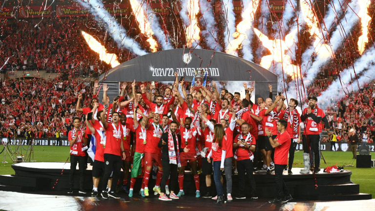 Voetbalteam in rode shirts viert kampioenschap op podium met trofee, vuurwerk en confetti in een vol stadion