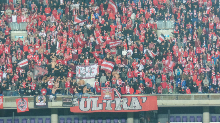 Volle voetbaltribune met supporters in rood die vlaggen zwaaien en een groot ULTRAS-spandoek ophangen