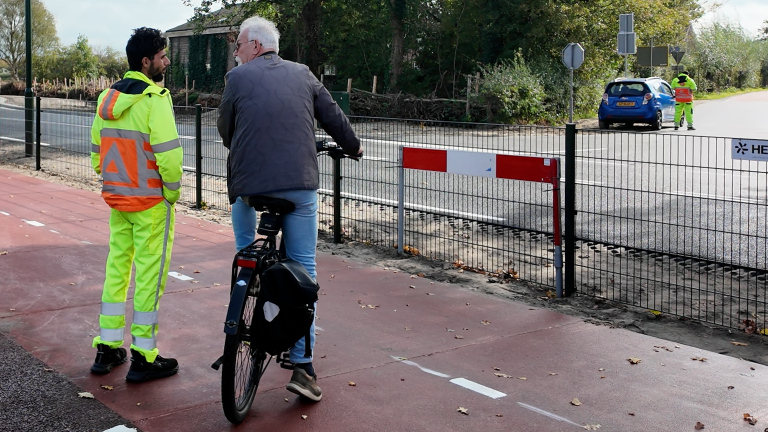 Wegwerker in fluorescerend pak praat met oudere fietser op fietspad naast weg, met auto en collega op de achtergrond