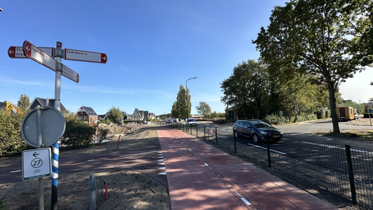 Wegwijzer met Egmond 6 en Limmen 2 bij rood fietspad langs een weg met auto’s, bomen en huizen onder blauwe lucht
