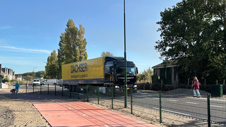 Grote gele vrachtwagen met opschrift DACHSER rijdt door woonwijk langs rood fietspad en groen hekwerk