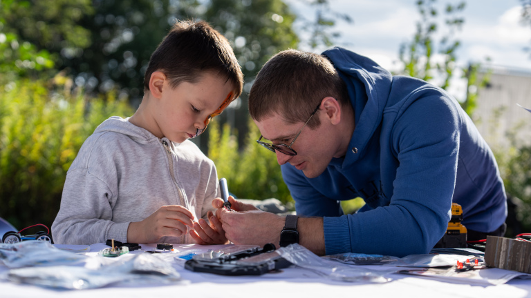 Man helpt kind met solderen aan een klein elektronica-onderdeel aan een tafel buiten
