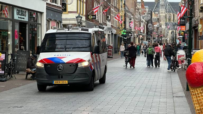 Nederlandse politie-bus in een drukke winkelstraat met voetgangers, fietsen en rood-witte vlaggen en lichtbollen boven de straat