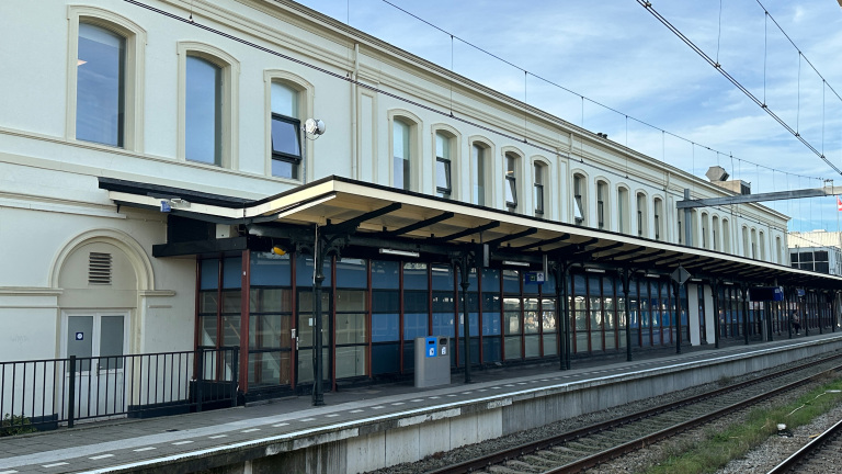 Lang perron met overkapping langs een wit stationsgebouw, met bovenleidingdraden en spoorrails op de voorgrond