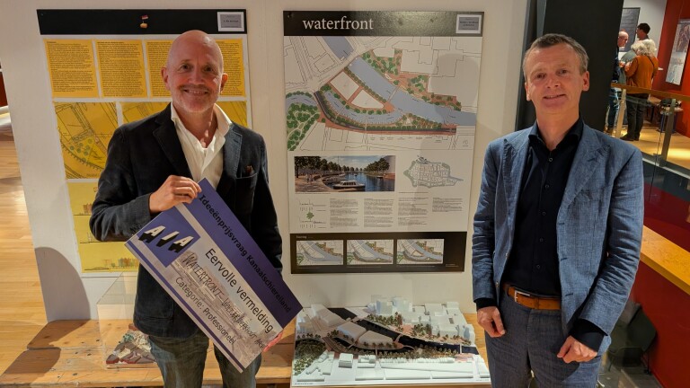Twee mannen bij een presentatiebord “waterfront”, met een maquette; één houdt een AIA-certificaat vast