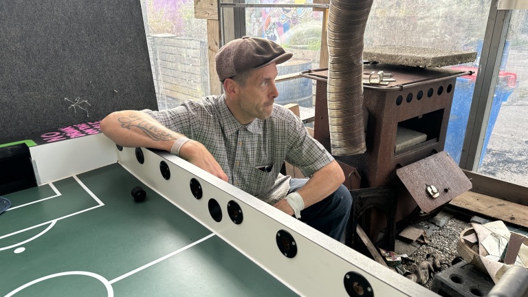 Man met pet en tatoeage leunt op een tafelvoetbaltafel naast een roestige houtkachel in een rommelige ruimte