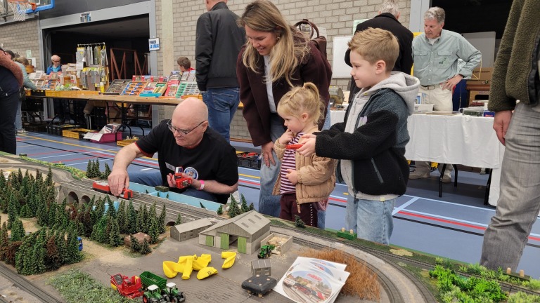 Man laat kinderen een modeltreinbaan bedienen op een beurs, met miniatuurlandschap en tafels met spullen op de achtergrond