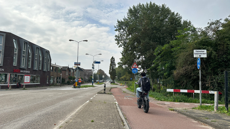 Fietser met rugzak op rood fietspad langs een weg met winkels en bomen, bij een bord richting Castricummerwerf