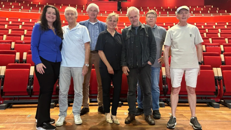 Zeven volwassenen poseren in een auditorium voor rijen rode stoelen