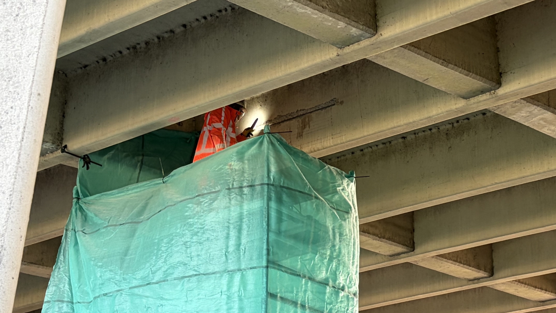 Bouwvakker in oranje hesje werkt onder een betonnen brug, achter groen afdekzeil op steiger