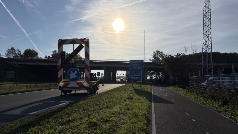 Pijlwagen met linksafpijl bij een viaduct en wegwijzer naar Jachthavens, Zaanstad en Amsterdam, met laagstaande zon