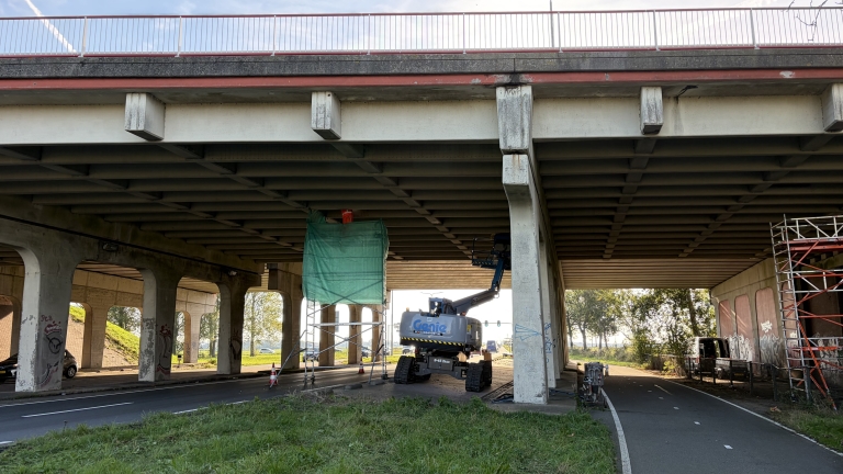 Werkzaamheden onder een viaduct met hoogwerker en steiger met groen net, naast fietspad en pijlers met graffiti