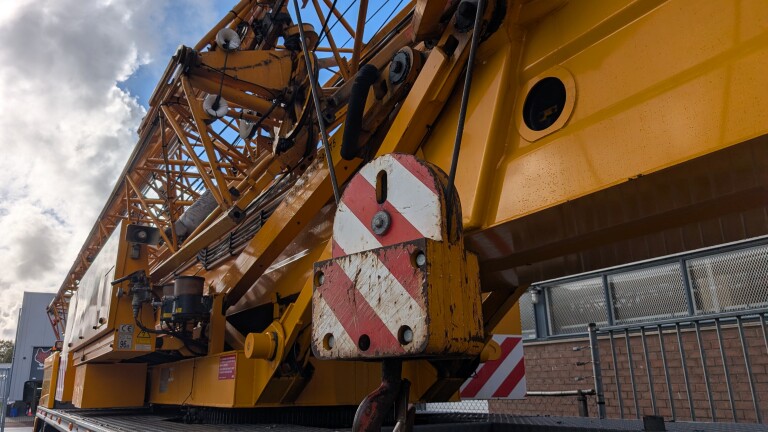 Detail van een gele hijskraan met hijshaak en rood-wit waarschuwingsbord, tegen een bewolkte lucht