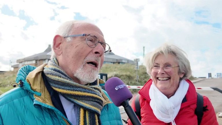 Oudere man met sjaal spreekt in microfoon terwijl vrouw naast hem lacht, buiten bij duinen onder bewolkte lucht