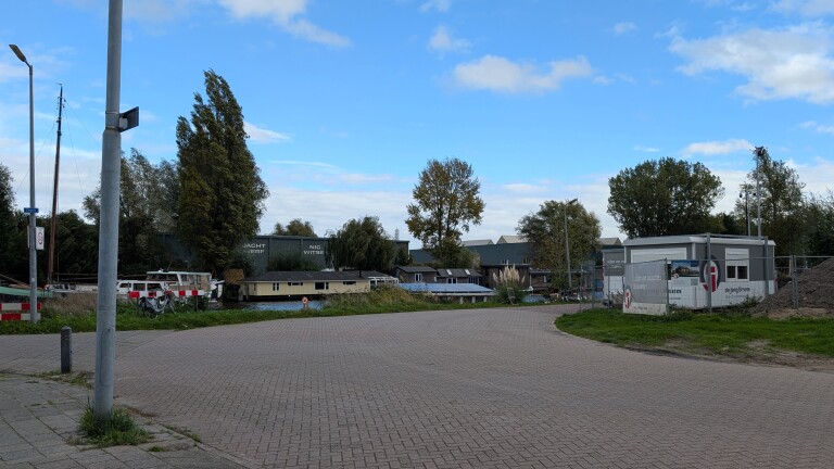 Lege weg langs een kanaal met woonboten, bomen en een bouwplaats met keet en hekwerk