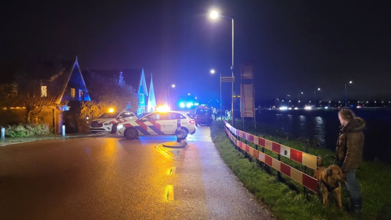 Politieauto’s met knipperlichten blokkeren ’s nachts de weg langs een kanaal; toeschouwer met hond staat rechts