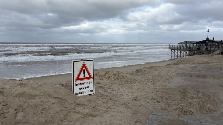Waarschuwingsbord “Instortingsgevaar zandduinen” op winderig strand met hoge golven en strandpaviljoen