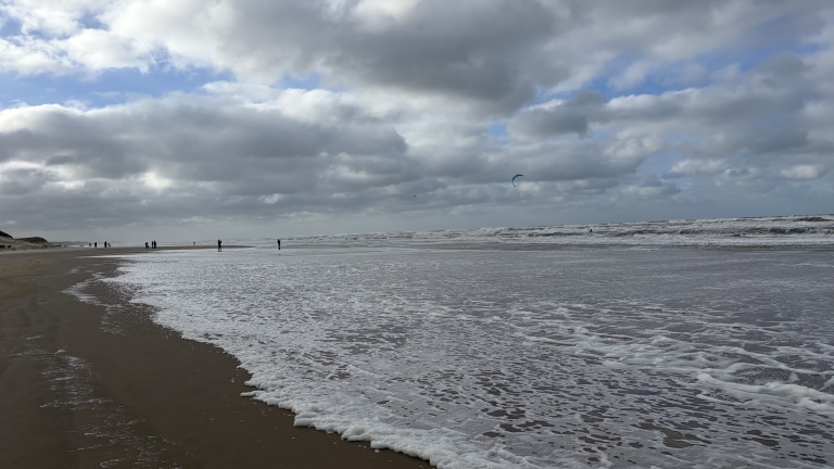 Wijd strand met schuimende branding en enkele wandelaars, onder een bewolkte lucht met een kitesurfvlieger in de verte