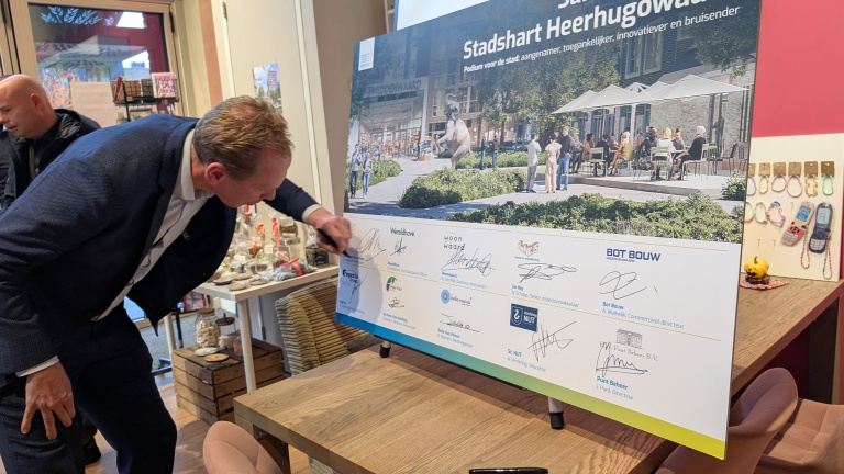 Man zet zijn handtekening op een groot presentatiebord over Stadshart Heerhugowaard, met meerdere logo’s en signatures