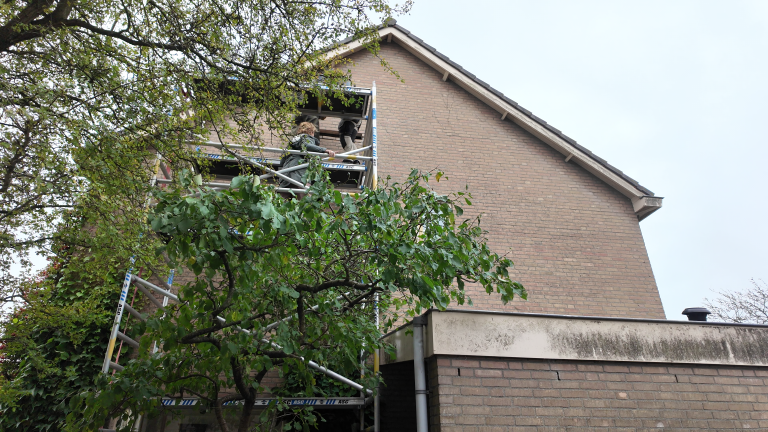 Twee mensen op steiger bij een raamopening in de gevel van een bakstenen huis, deels verscholen achter bomen