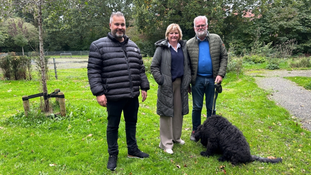 Drie personen poseren in een groen park; één houdt een hondenriem vast en een zwarte hond zit op de voorgrond