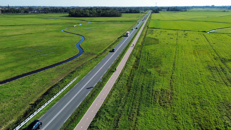 Luchtfoto van een lange weg met fietspad langs groene weilanden en een slingerende sloot, met auto’s en fietsers