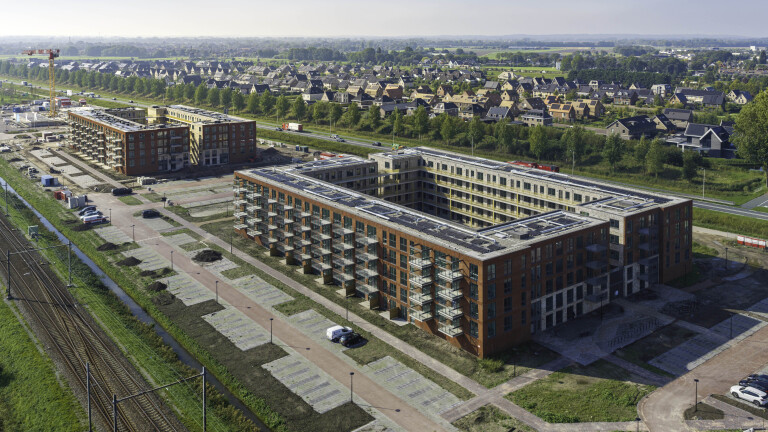 Luchtfoto van nieuwbouwappartementen met balkons naast spoorlijn, met woonwijk en groen op de achtergrond