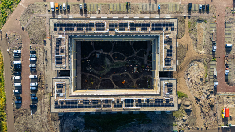Luchtfoto van vierkant gebouw in aanbouw met zonnepanelen op het dak en een open binnenplaats vol bouwgrond
