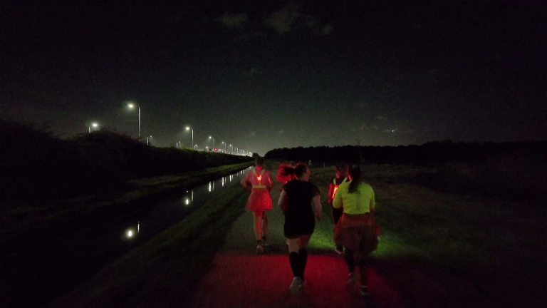 Groep hardlopers met rode lampjes op een donker pad langs een kanaal, met straatverlichting in de verte