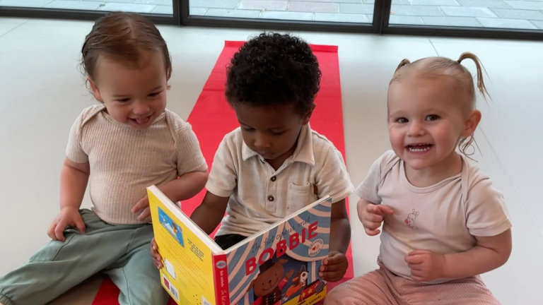Drie peuters zitten op een rode mat; één leest een prentenboek, de twee anderen kijken lachend mee