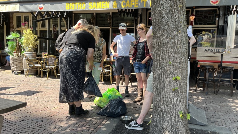 Mensen verzamelen zich rond zakken afval op een zonnige stoep voor een bistro en café genaamd 'Bistro Samen & Café'.