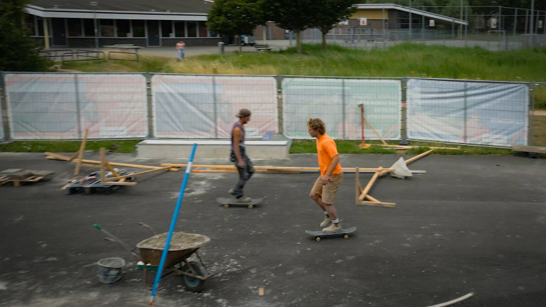 Twee skaters rijden over een asfaltterrein met verspreid hout en een kruiwagen, achter een bouwhek