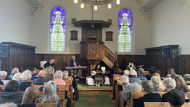 Mensen luisteren naar een muzikaal optreden in een kerk met kleurrijke glas-in-loodramen.