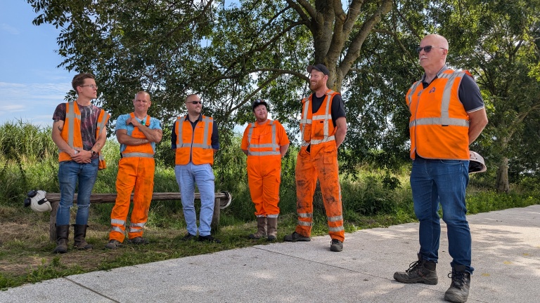 Midden in de natuur staan zes mannen in werkkleding en veiligheidsvesten samen te praten.