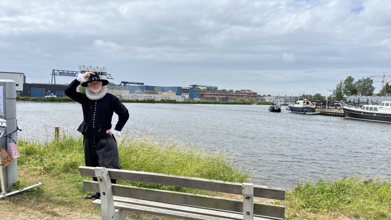 Een man in historische kleding poseert naast een bankje aan de oever van een rivier, met industrielandschap en boten op de achtergrond.
