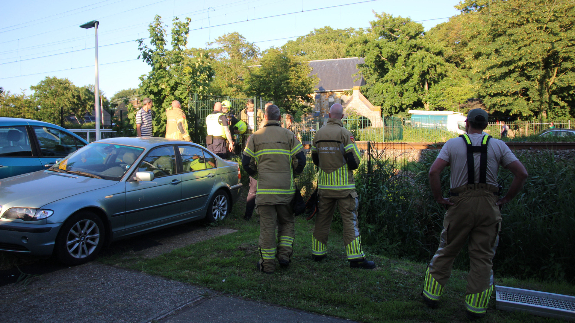 Brandweerlieden in uniform staan bij een geparkeerde auto en kijken naar een gebied omheind met groen. Op de achtergrond zijn bomen en een klein huisje zichtbaar.